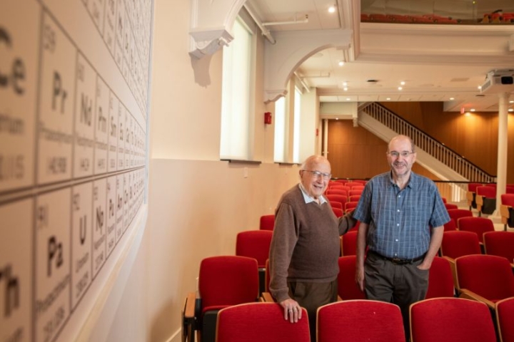 professor, alum stand in row of red chairs